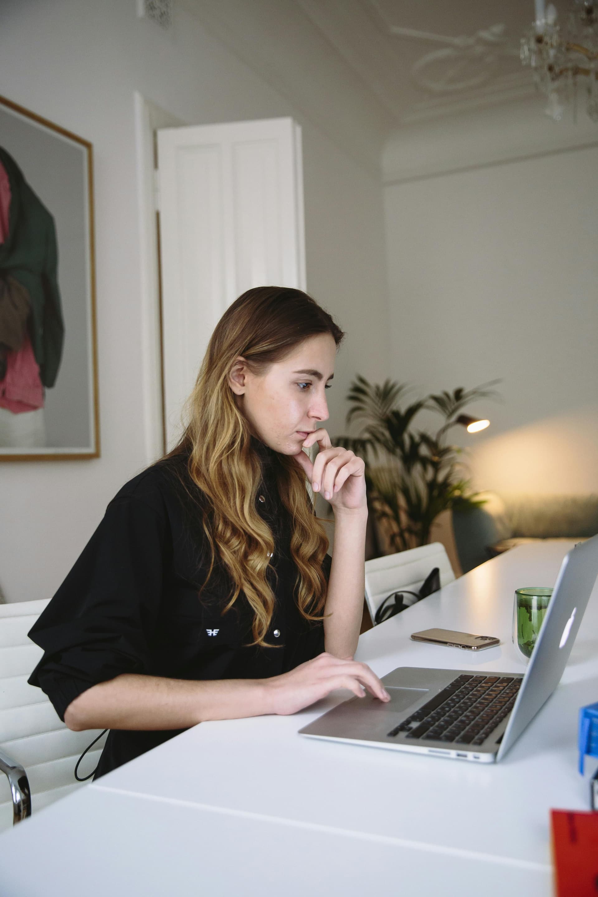 A person sits at a white desk working on a laptop, resting their chin on one hand while using the trackpad with the other. A smartphone and a glass sit nearby, with indoor plants and soft lighting in the background, creating a calm, focused workspace.