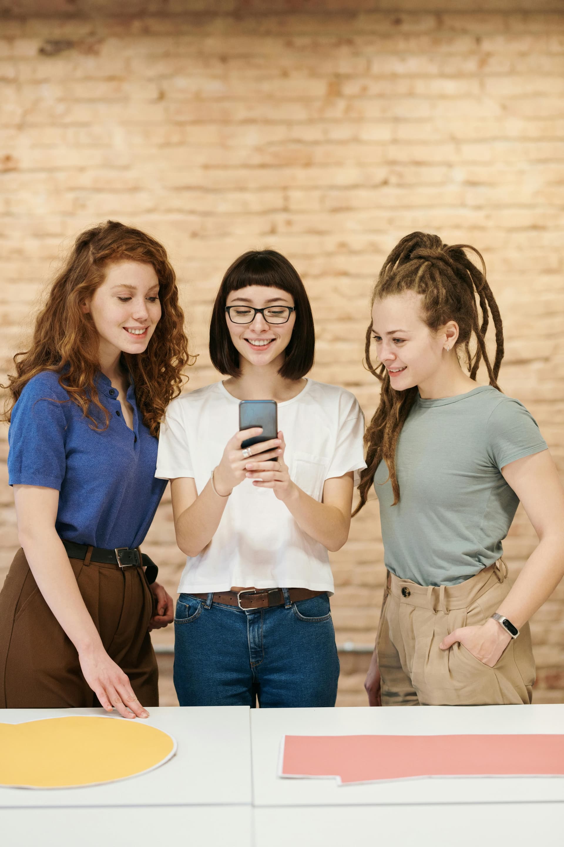 Three people stand together around a table, smiling and looking at a smartphone held by one of them. Color samples or papers lie on the table, with a brick wall in the background, suggesting a collaborative and creative discussion.